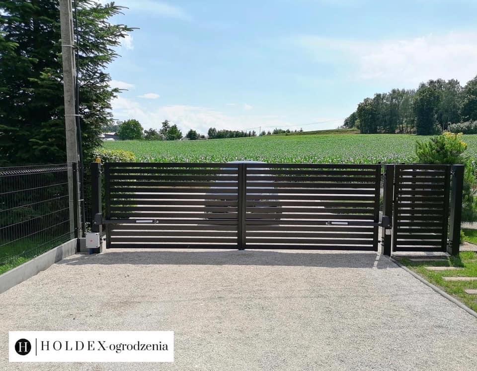 Nowoczesna, pozioma brama przesuwna w kolorze ciemnego brązu z automatyką, na tle zielonego pola i błękitnego nieba.