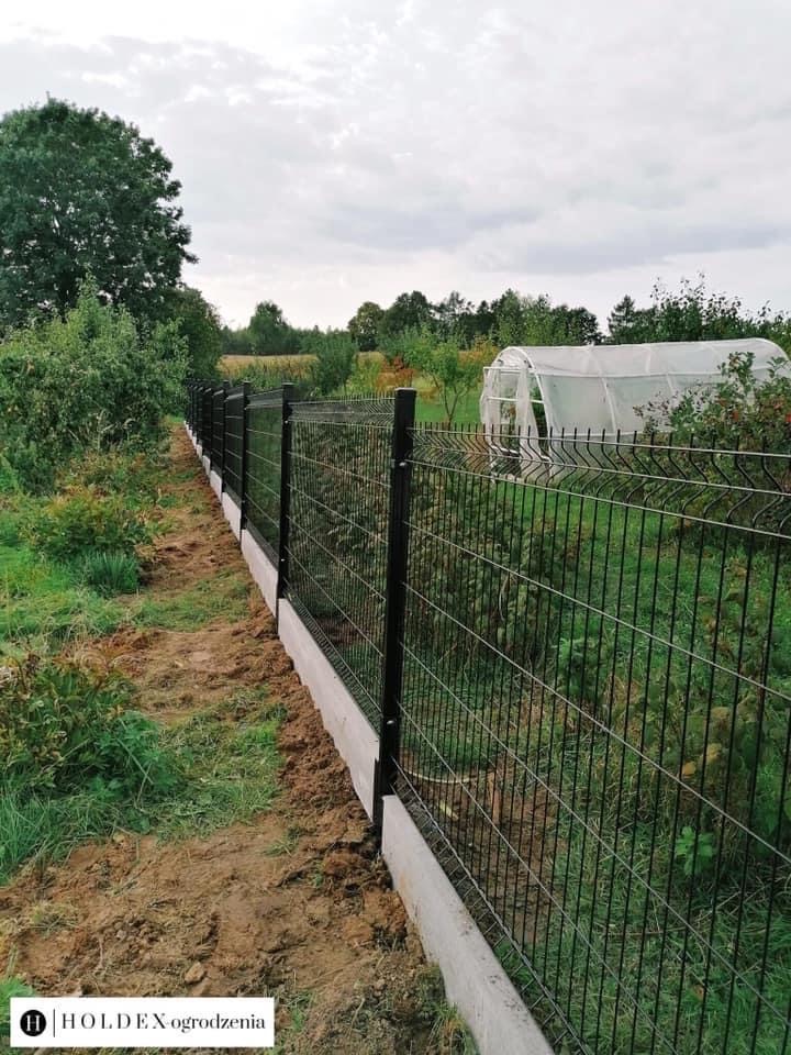 Nowoczesne ogrodzenie panelowe z podmurówką betonową, widok na ogród z tunelem foliowym w tle, ziemia świeżo naruszona po montażu.