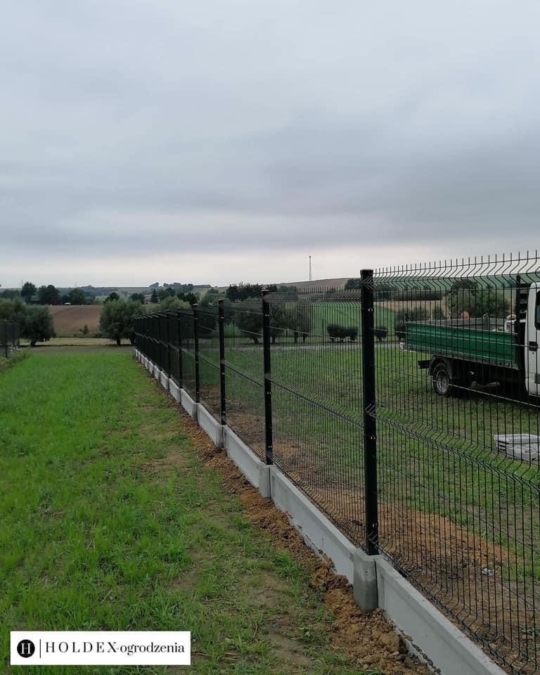 Panelowe ogrodzenie z podmurówką betonową, widok z boku na tle zielonego trawnika i pola, z samochodem dostawczym w tle. Logo firmy w lewym dolnym rogu.