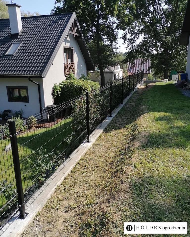 Nowoczesne, czarne ogrodzenie panelowe z podmurówką betonową, oddzielające posesję z domem jednorodzinnym i bujną zielenią, widok z perspektywy trawnika.