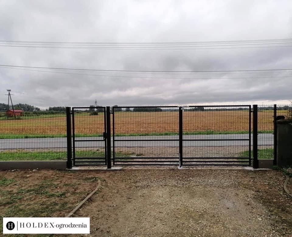 Czarna, metalowa brama dwuskrzydłowa z siatki panelowej, zamontowana na betonowym fundamencie, widok na polną drogę i pole w pochmurny dzień, logo Holdex-ogrodzenia w lewym dolnym rogu.