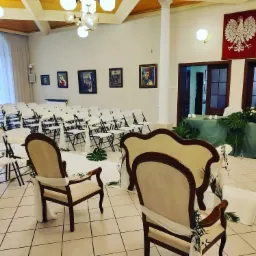 Sala przygotowana na uroczystość z rzędami białych krzeseł, dwa zdobione fotele na pierwszym planie, stół z zielonym obrusem i kwiatami w tle, godło Polski na ścianie.