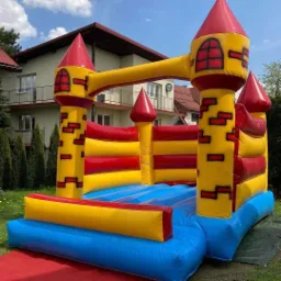 Kolorowy, dmuchany zamek w stylu średniowiecznym na trawie, z czerwonym dywanem u wejścia, na tle domu z balkonem.