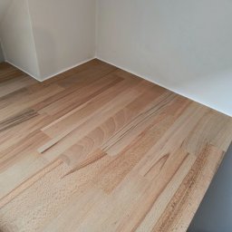 Mateusz - Blat drewniany z klejonego drewna bukowego, zamontowany w rogu pomieszczenia z białymi ścianami, widoczne detale struktury drewna i łączenia klejonego.