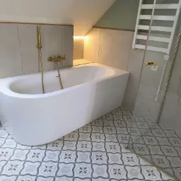 Nowoczesna biała wanna wolnostojąca z złotą armaturą, umieszczona w łazience z szaro-beżowymi płytkami ściennymi i dekoracyjną podłogą w kwiatowe wzory. Widoczny grzejnik drabinkowy za szklaną...