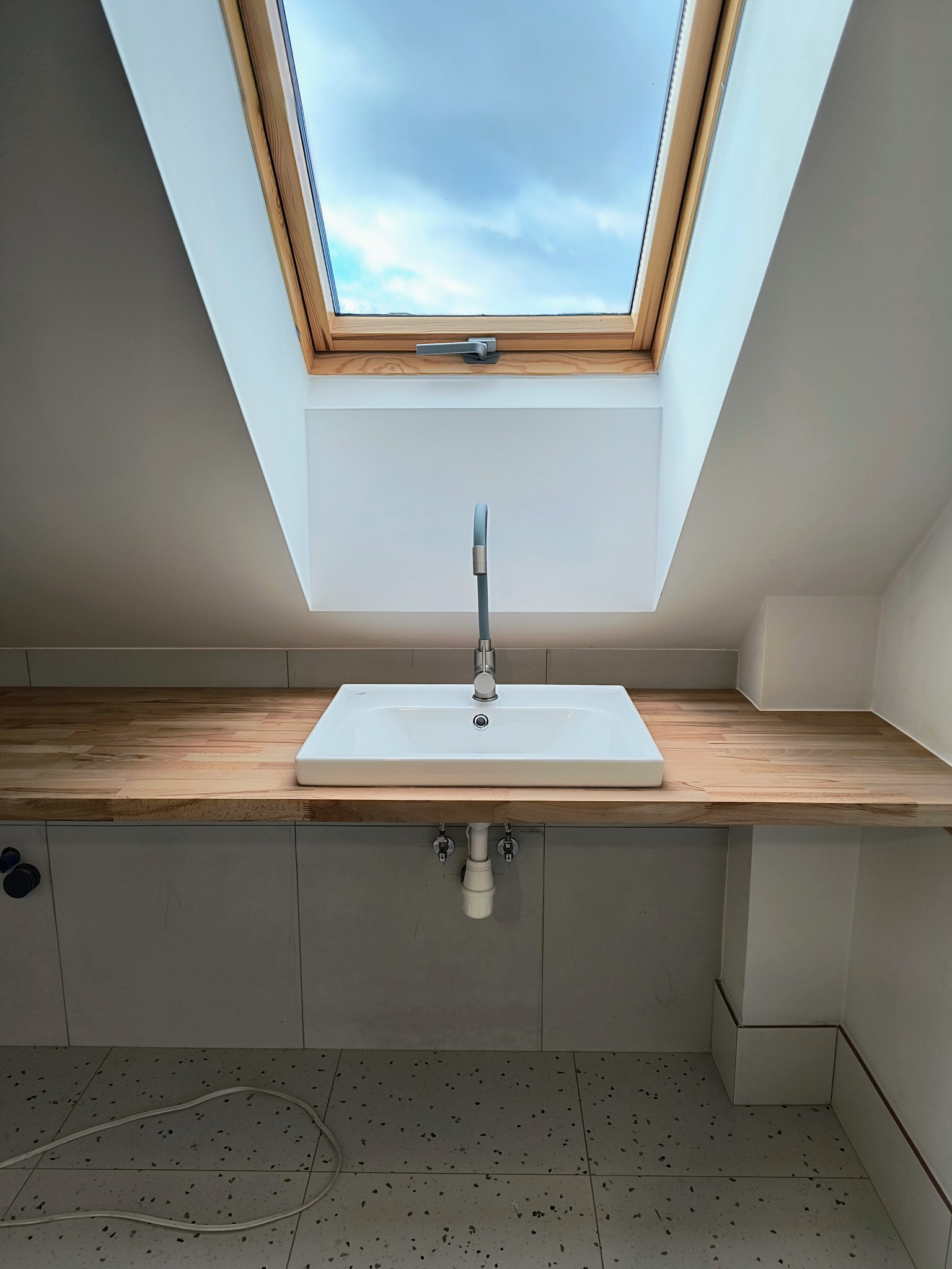 Biała, prostokątna umywalka z chromowaną baterią zamontowana w drewnianym blacie pod oknem dachowym w łazience wykończonej szarymi płytkami.