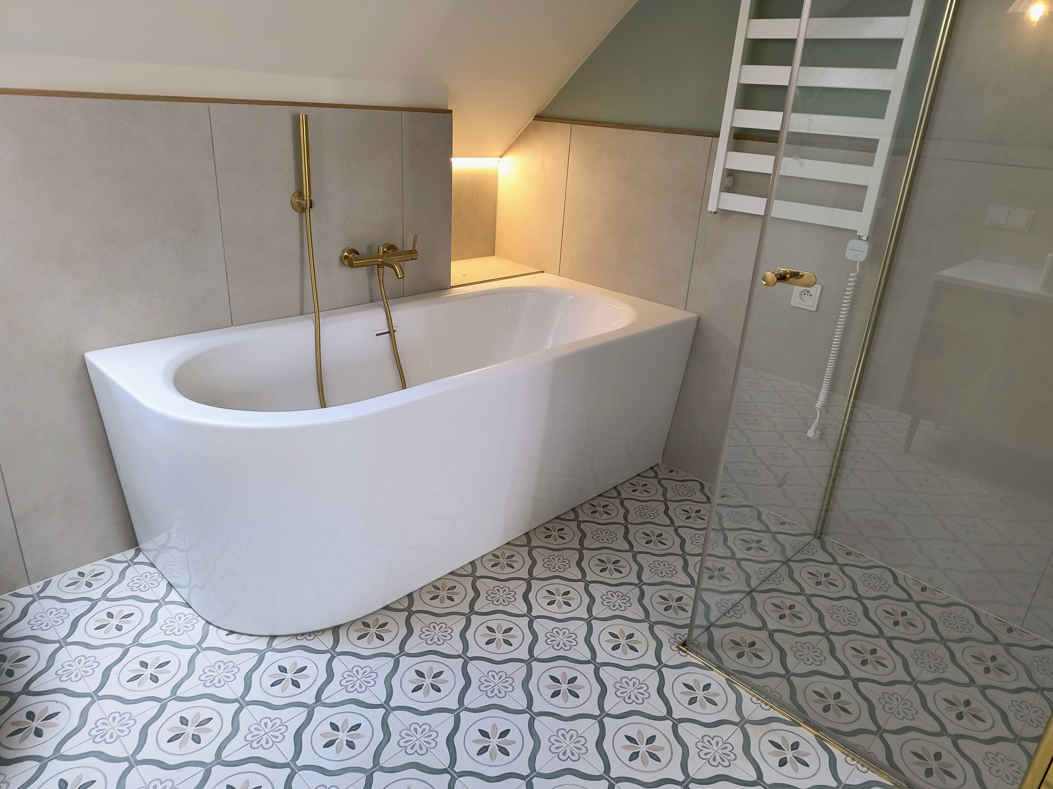 Nowoczesna biała wanna wolnostojąca z złotą armaturą, umieszczona w łazience z szaro-beżowymi płytkami ściennymi i dekoracyjną podłogą w kwiatowe wzory. Widoczny grzejnik drabinkowy za szklaną...