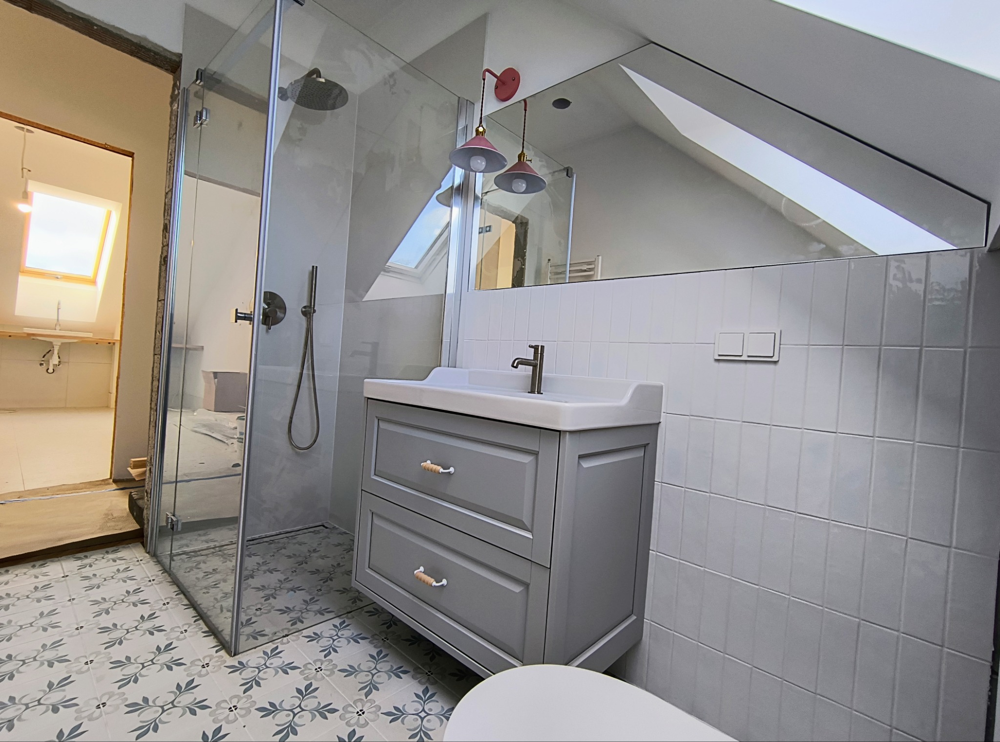 Ukończona łazienka na poddaszu ze szklaną kabiną prysznicową, szarą szafką z umywalką i ozdobnymi płytkami podłogowymi.