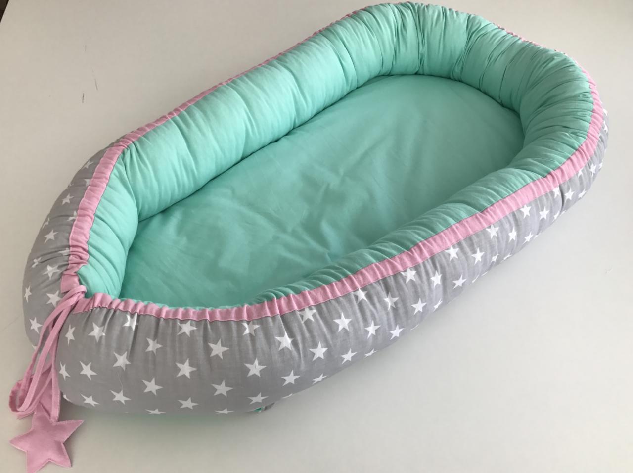 Miękkie gniazdko niemowlęce w kolorze szarym w białe gwiazdki, z miętowym wnętrzem i różowymi akcentami, leżące na białym tle.