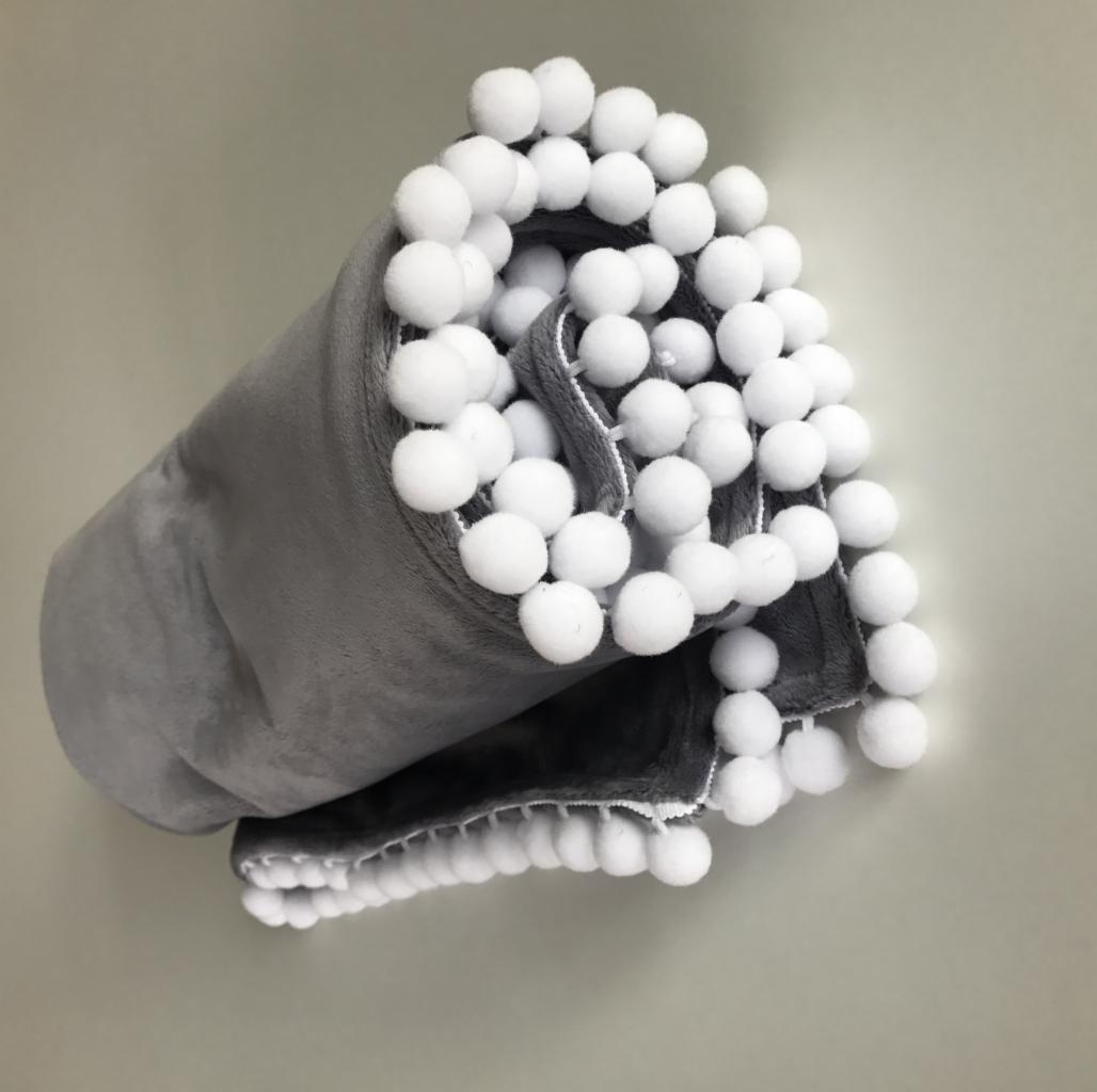 Zrolowany szary koc z miękkiego pluszu, ozdobiony rzędem białych pomponów na krawędzi, widoczny z bliska na jednolitym tle.