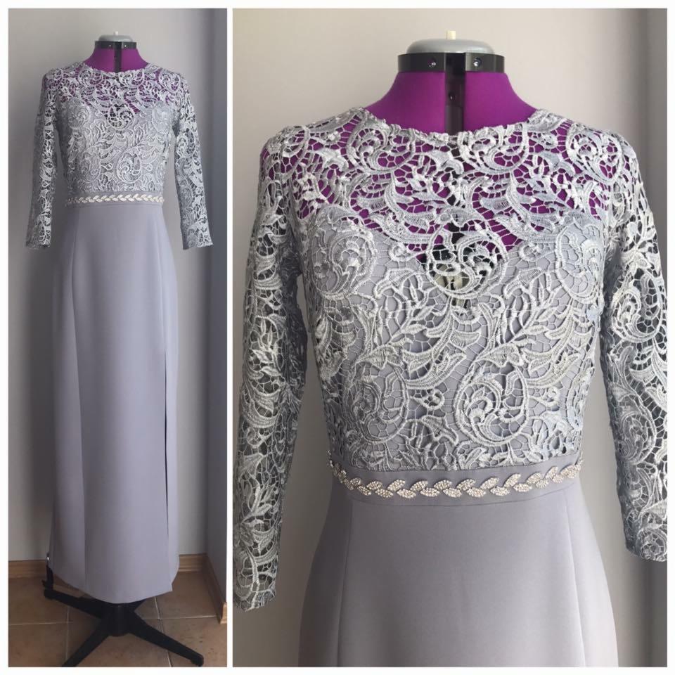 Długa, szara sukienka z koronkową górą i długimi rękawami, ozdobiona delikatnym paskiem z motywem liści, prezentowana na fioletowym manekinie.