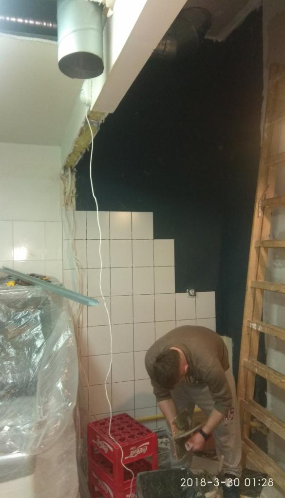 Układanie białych płytek na ścianie w trakcie remontu łazienki, widoczna drabina i skrzynki Coca-Coli, fragment instalacji wentylacyjnej w suficie.