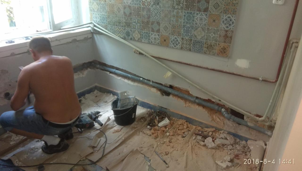 Mężczyzna w trakcie skuwania ściany w łazience, widoczne instalacje wodno-kanalizacyjne, narzędzia i gruz na podłodze.