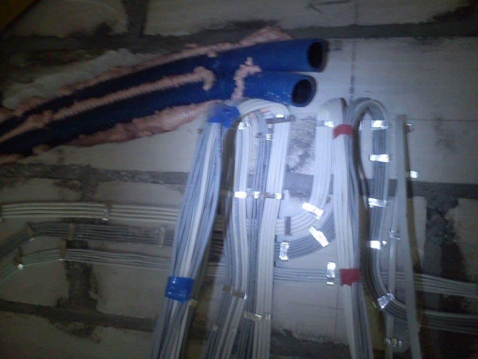 Ściana z widocznymi przewodami elektrycznymi w peszlach i kablami przymocowanymi klamrami, w trakcie instalacji. Widoczne fragmenty muru i niebieskie rury ochronne.
