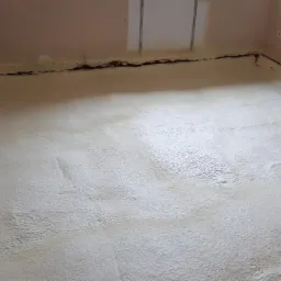 Podłoga w trakcie ocieplania pianką PUR, widoczne nier&oacute;wności pianki oraz fragmenty ściany z elementami instalacji.