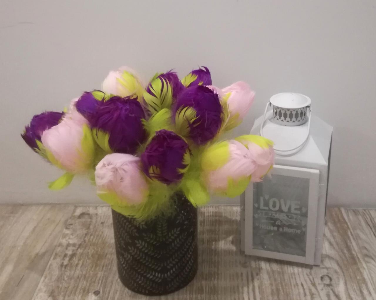 Dekoracyjny bukiet z piór w odcieniach fioletu, różu i limonki, umieszczony w ciemnym wazonie z wzorem jodełki, obok białego lampionu z napisem 'LOVE' na drewnianym blacie.