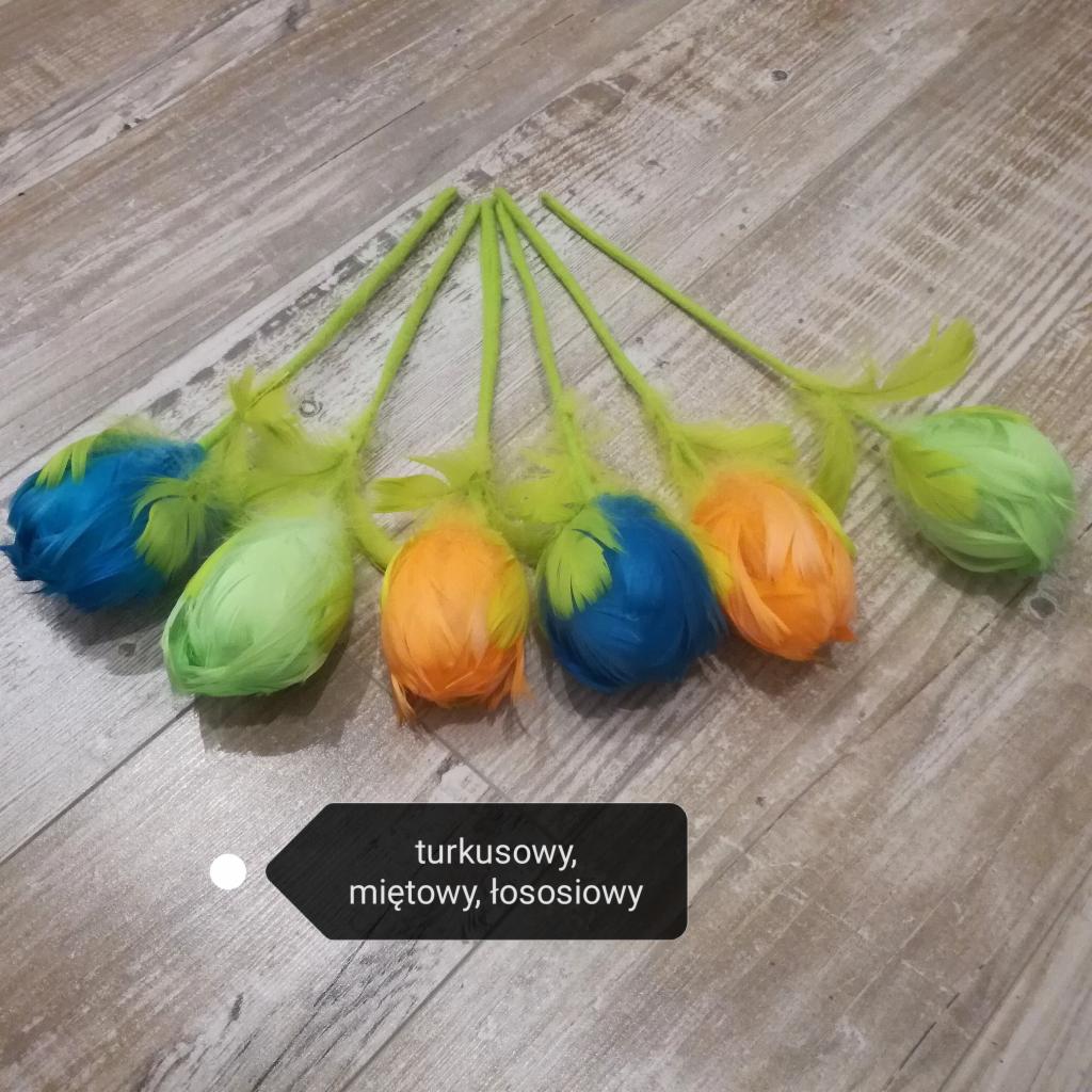 Sześć dekoracyjnych, kolorowych jajek z piórkami na zielonych łodygach, ułożonych na drewnianym tle.