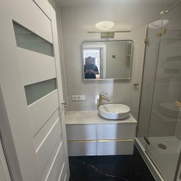 Grupa prace wykończeniowe - Elegancka łazienka po remoncie: czarna marmurowa podłoga, biała szafka z umywalką i złotymi akcentami, szklana kabina prysznicowa.