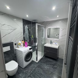 Grupa prace wykończeniowe - Nowoczesna łazienka w odcieniach szarości z marmurowymi płytkami, kabiną prysznicową i pralką. Oświetlenie punktowe podkreśla elegancję wnętrza.