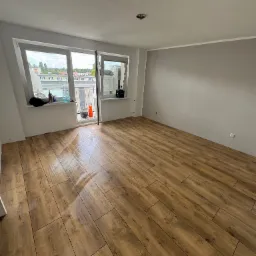 Jasne panele podłogowe imitujące drewno w pustym pokoju z szarą ścianą i oknem balkonowym. Widok na zewnątrz.