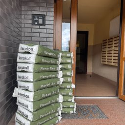 Grupa prace wykończeniowe - Stos worków z zaprawą Kerakoll Planogel Ultra przy wejściu do budynku z szarą cegłą. Widok na klatkę schodową i skrzynki pocztowe.