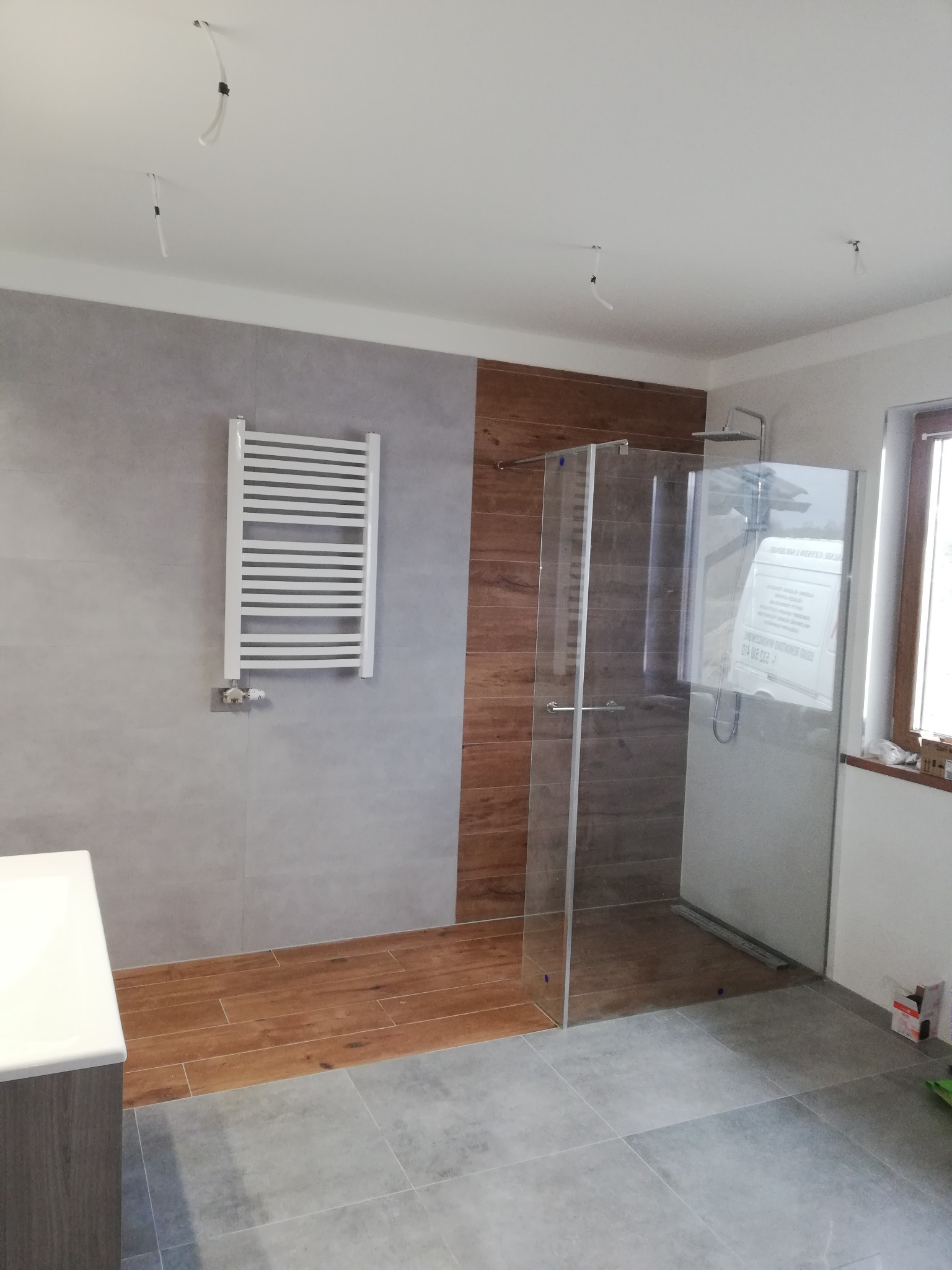 Nowoczesna łazienka z kabiną prysznicową ze szklanymi drzwiami, grzejnikiem drabinkowym na szarej ścianie, drewnopodobnymi panelami ściennymi i szarą podłogą z płytek; widoczne niedokończone...