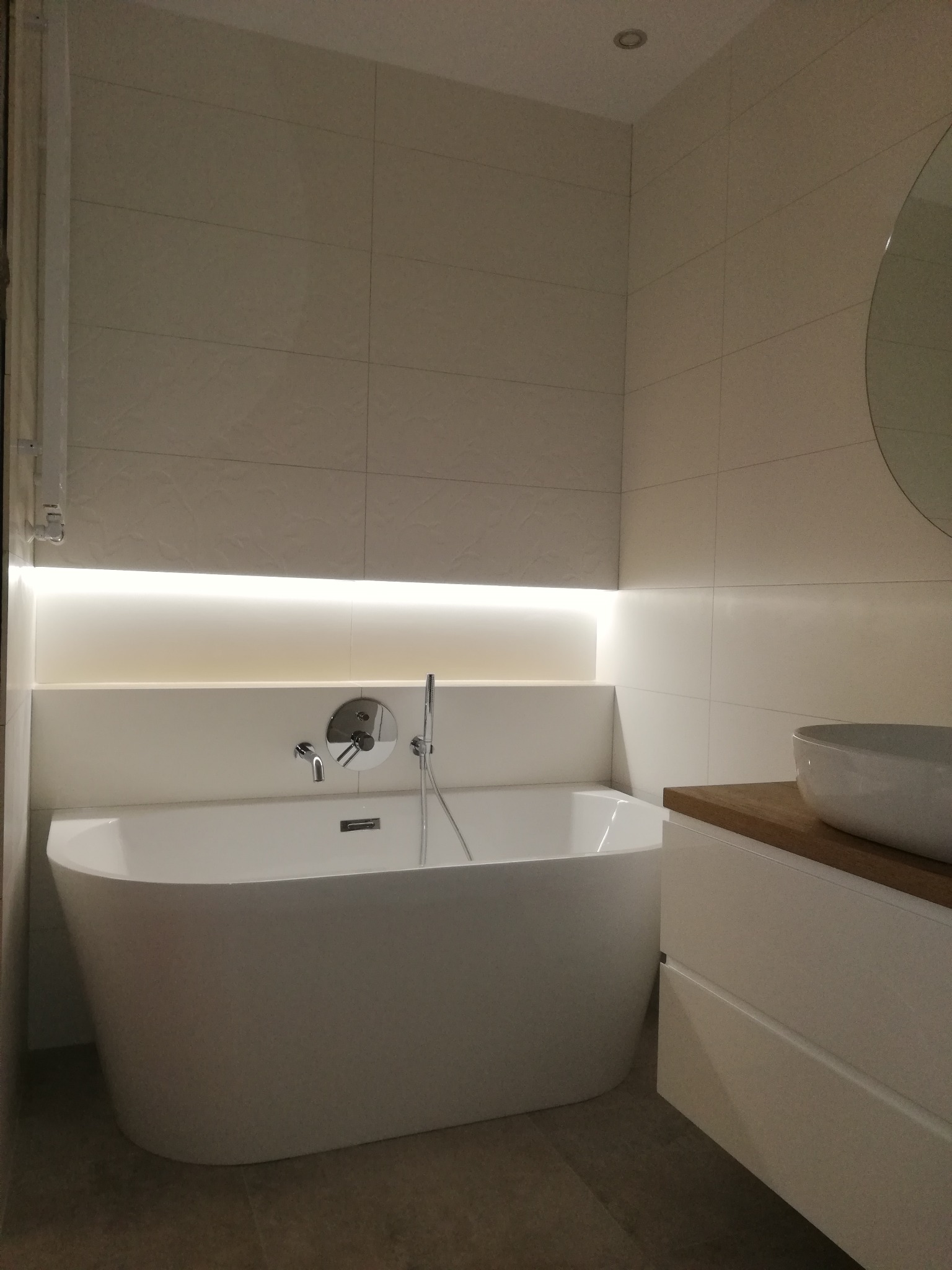 Nowoczesna łazienka z wolnostojącą białą wanną, podświetleniem LED nad wanną, beżowymi płytkami na ścianach i umywalką nablatową na drewnianym blacie.