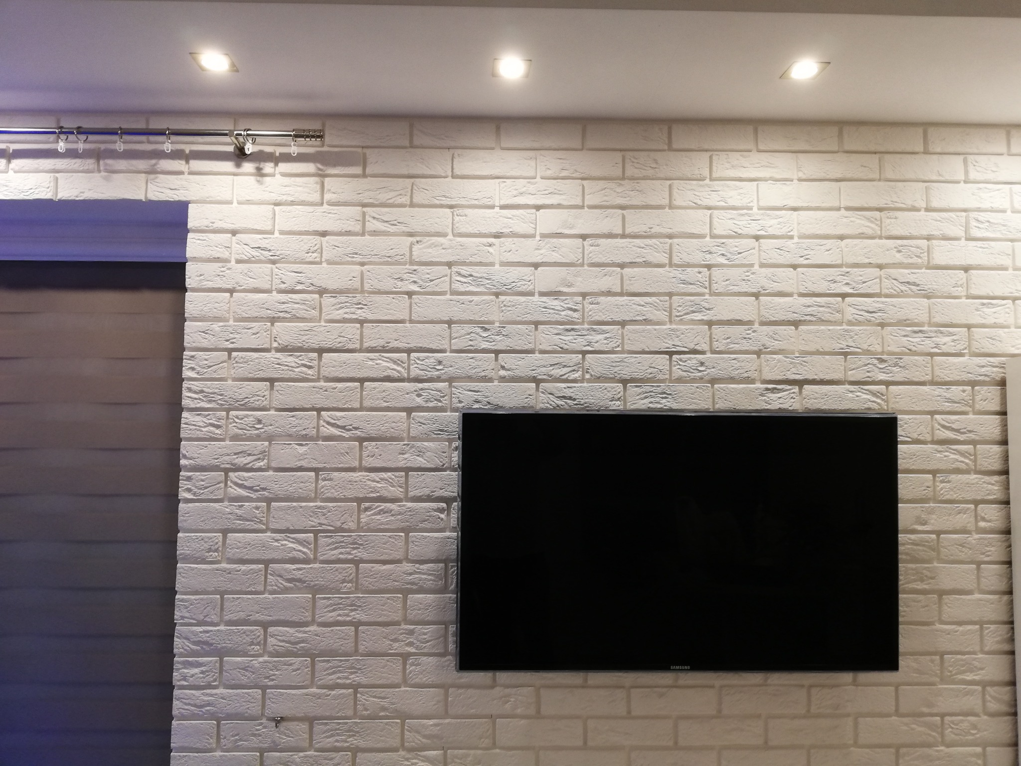 Ściana z białej cegły dekoracyjnej z zamontowanym telewizorem i karniszem, oświetlenie punktowe sufitowe.