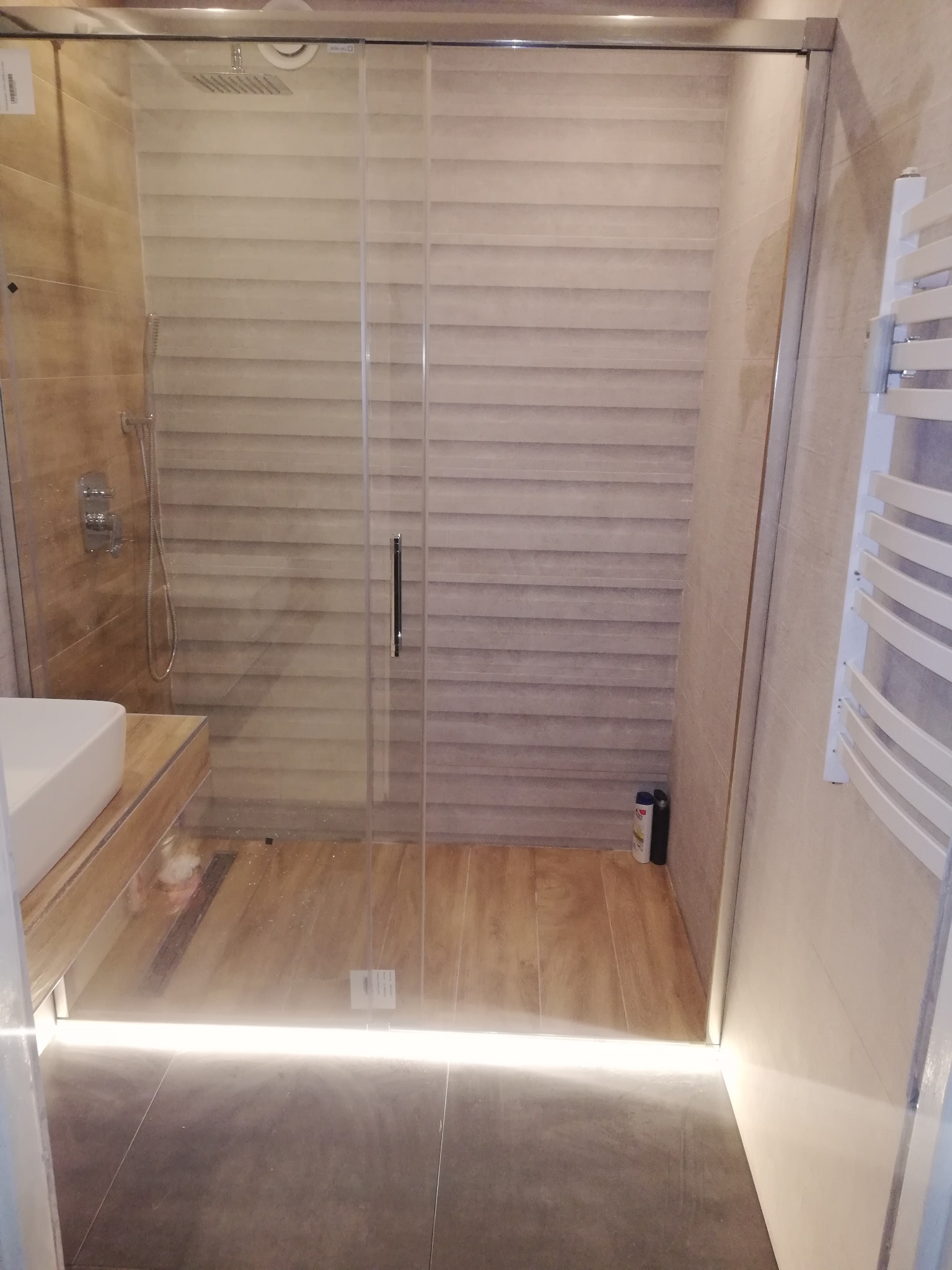 Nowoczesna kabina prysznicowa walk-in z odpływem liniowym, oświetleniem LED u dołu szyby i brązowymi płytkami imitującymi drewno na ścianach i podłodze.