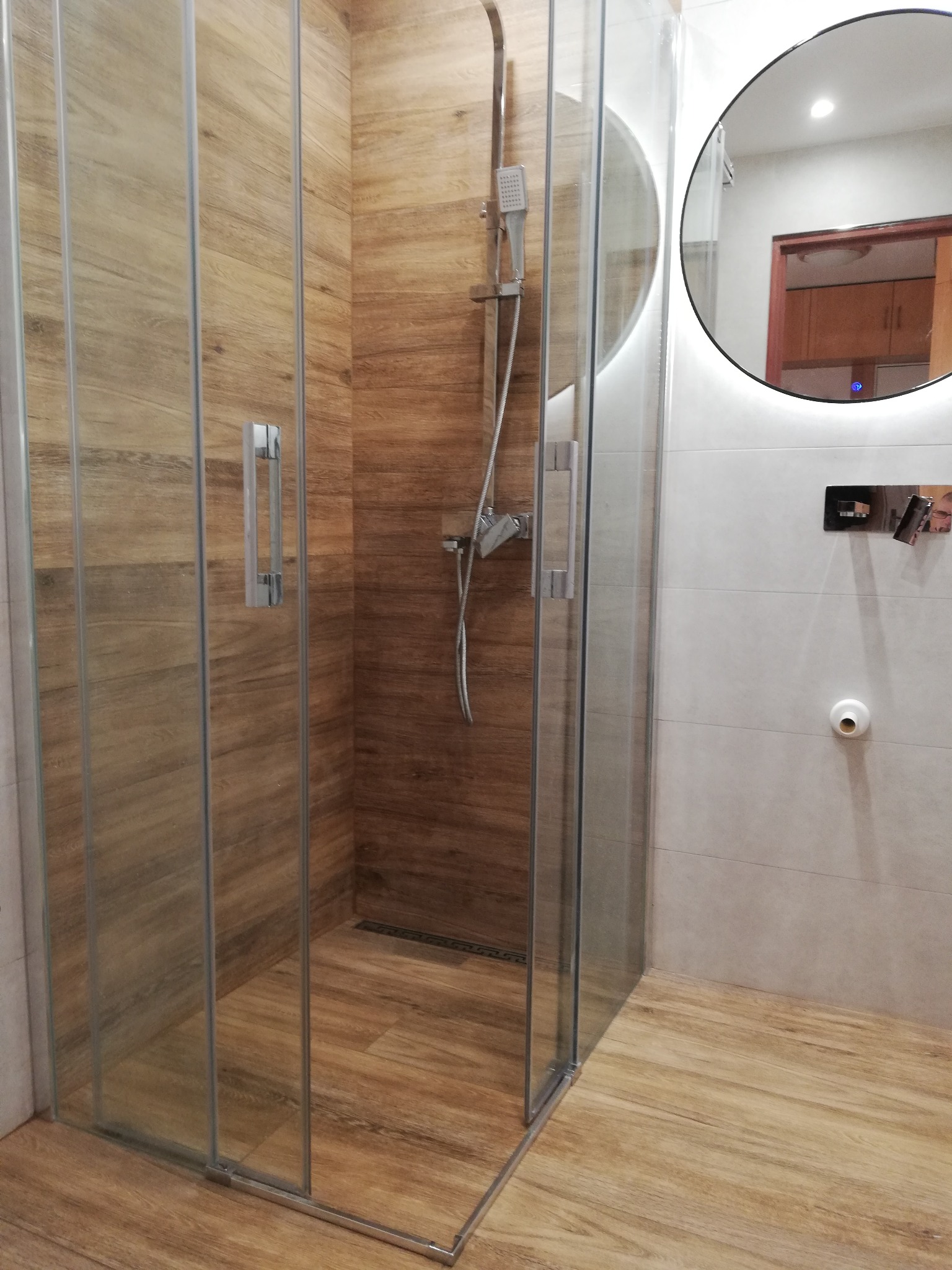 Nowoczesna kabina prysznicowa z przesuwnymi szklanymi drzwiami, wykończona płytkami imitującymi drewno, z okrągłym lustrem z podświetleniem LED w tle.
