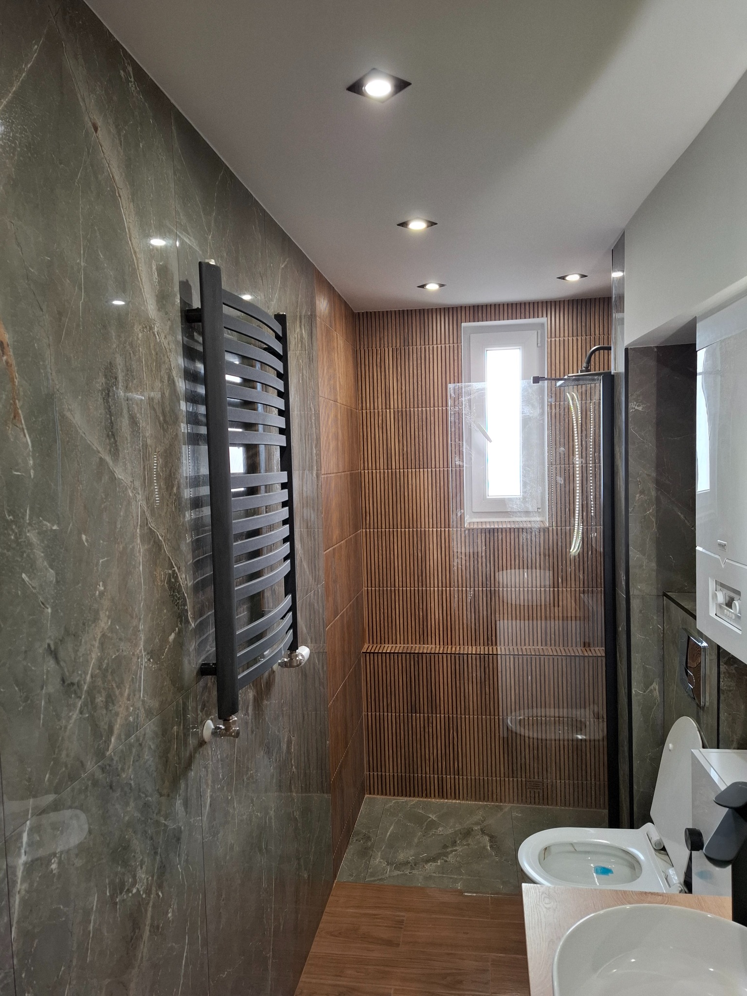Nowoczesna łazienka z marmurowymi płytkami, grzejnikiem drabinkowym i kabiną prysznicową z drewnianymi panelami w tle.