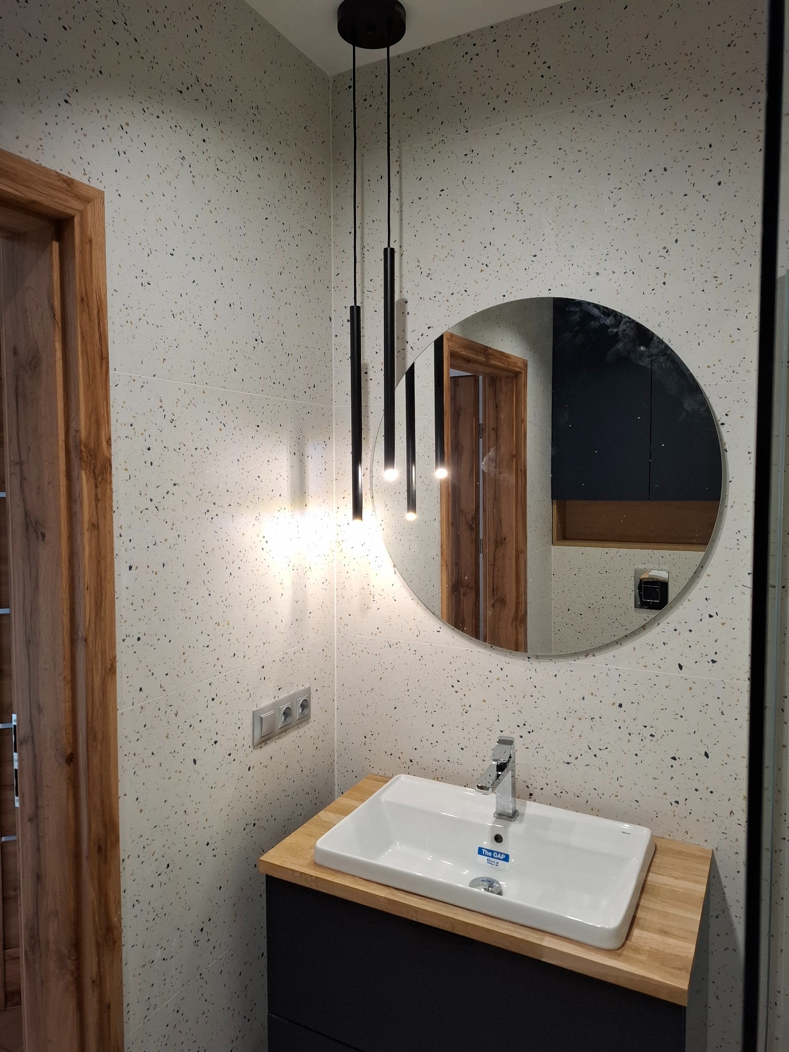 Nowoczesna łazienka z umywalką Roca na drewnianym blacie, okrągłym lustrem odbijającym fragment drzwi i czarnymi lampami wiszącymi na tle ściany wykończonej płytkami imitującymi lastryko.