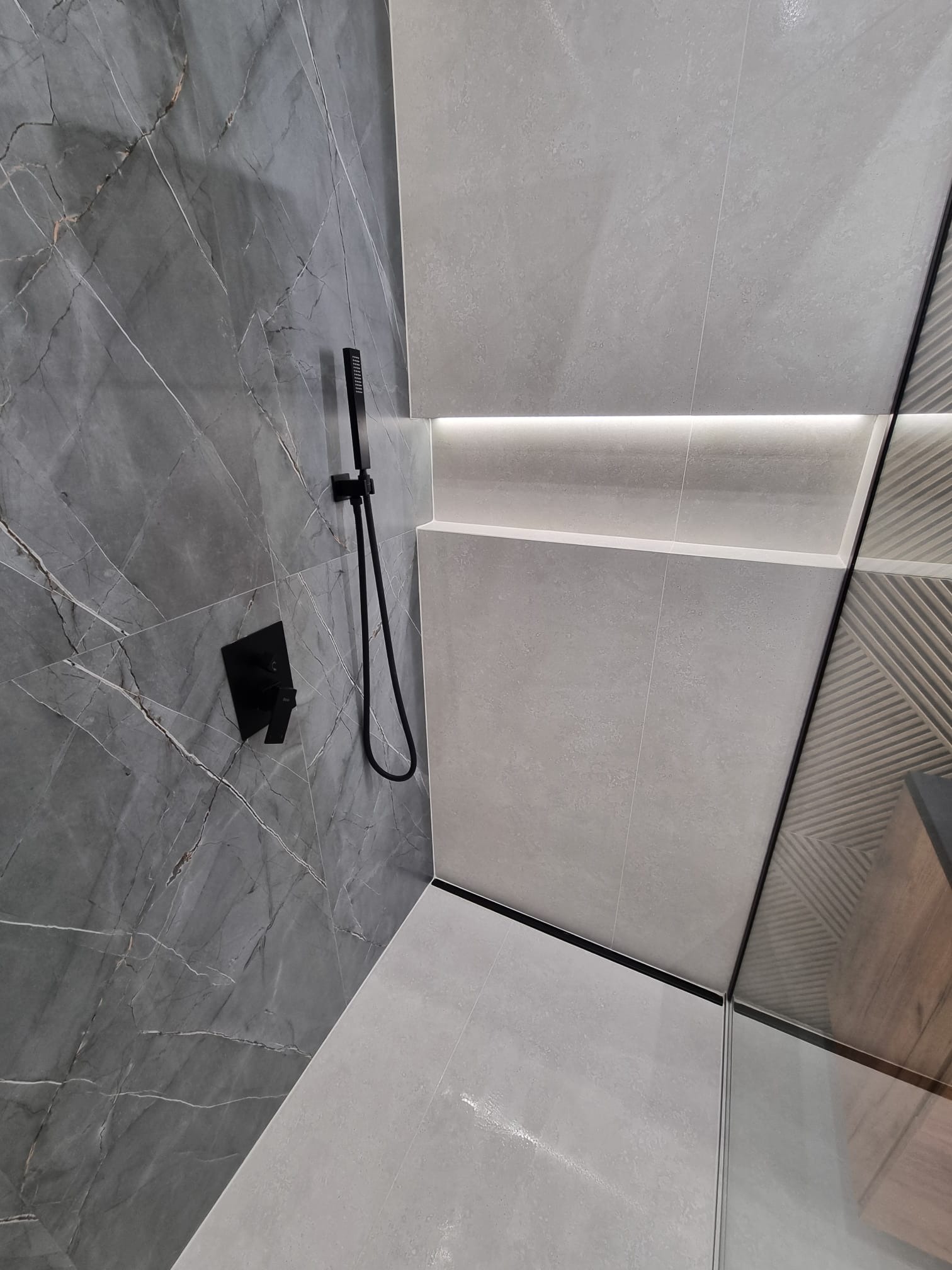 Nowoczesna kabina prysznicowa z szarą marmurową ścianą, czarną armaturą i subtelnym oświetleniem LED w półce.