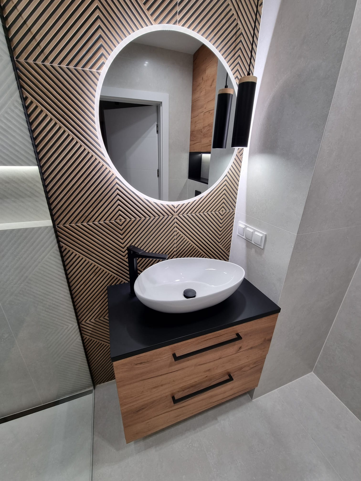 Nowoczesna łazienka z umywalką nablatową, szafką z drewnopodobnym fornirem i czarnymi uchwytami, okrągłym lustrem z podświetleniem LED oraz dekoracyjną ścianą z geometrycznym wzorem z drewna.