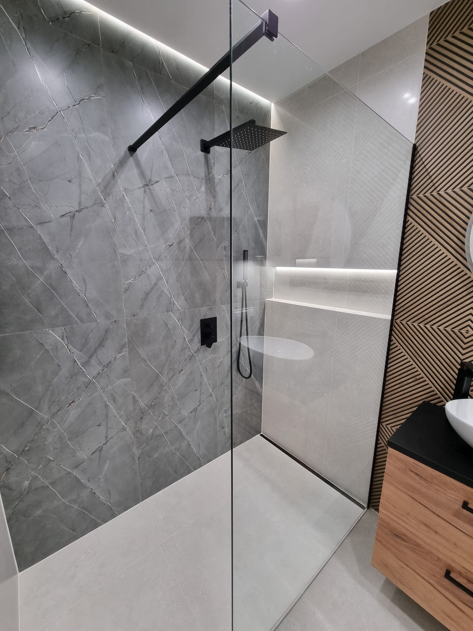 Nowoczesna kabina prysznicowa walk-in z czarną armaturą, szklaną ścianką i marmurową okładziną ścian, uzupełniona drewnianą ścianą z geometrycznym wzorem i umywalką nablatową.