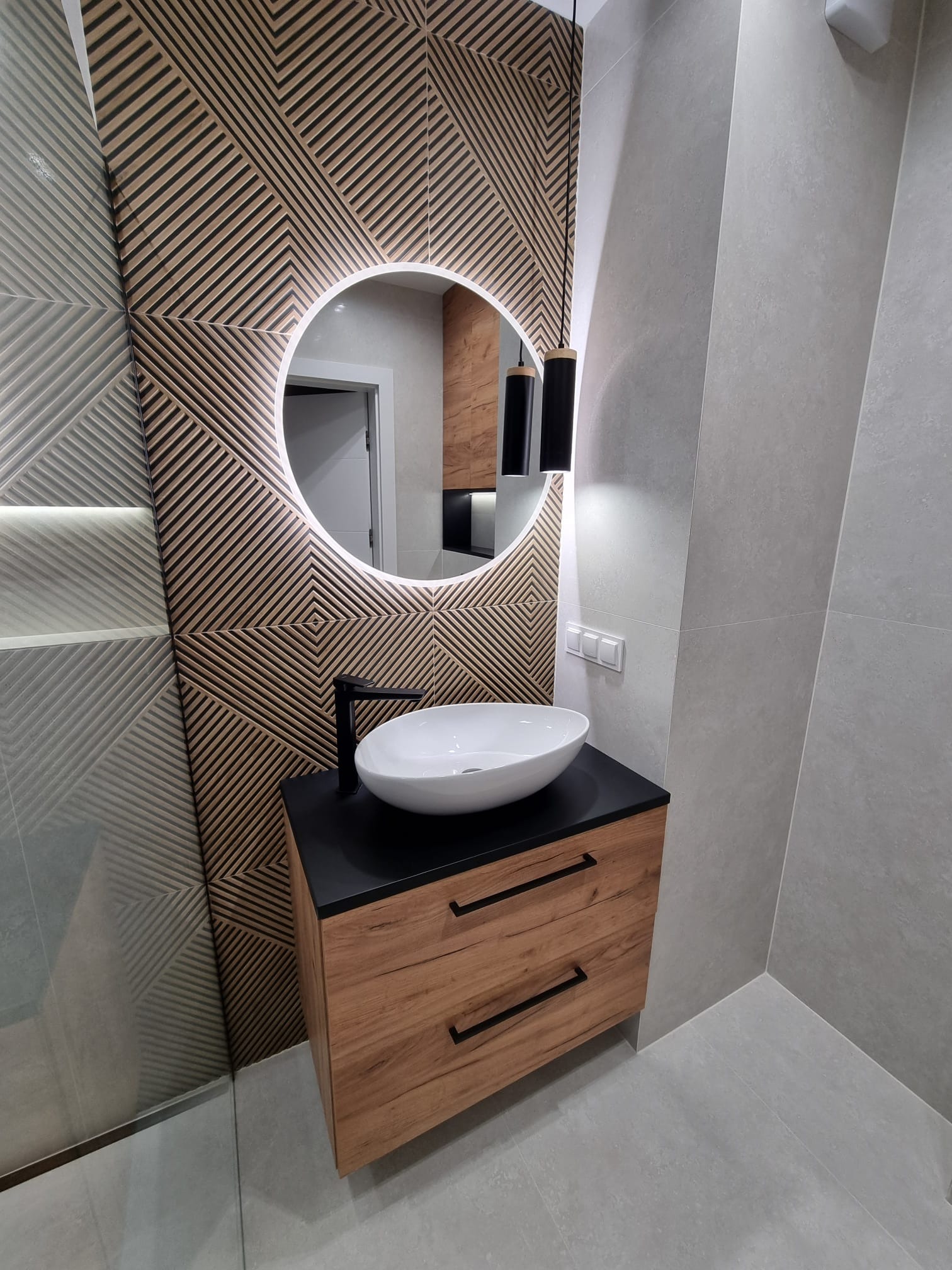 Nowoczesna łazienka z umywalką nablatową, szafką z drewnopodobnym frontem, okrągłym lustrem z podświetleniem LED i dekoracyjną ścianą z drewnianych paneli 3D.