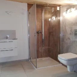 Nowoczesna łazienka po remoncie: kabina prysznicowa ze szklanymi drzwiami, podwieszana toaleta, biała szafka z umywalką i szara płytka imitująca beton na ścianach.