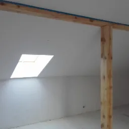 Poddasze z oknem dachowym i drewnianą konstrukcją nośną, ściany przygotowane do malowania, widoczne surowe drewno belek.