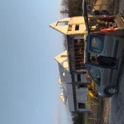 Wznoszenie ścian budynku z bloczków, widoczne szalunki i pręty zbrojeniowe, na pierwszym planie samochód dostawczy z otwartą klapą bagażnika i widocznym wyposażeniem budowlanym.