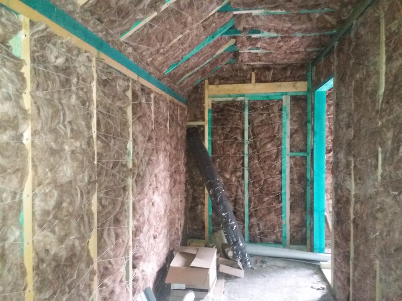 Wnętrze poddasza w trakcie prac: ściany i skośny sufit wypełnione wełną mineralną, widoczne drewniane konstrukcje szkieletowe pomalowane na zielono, kartony na podłodze i czarny rękaw wentylacyjny.