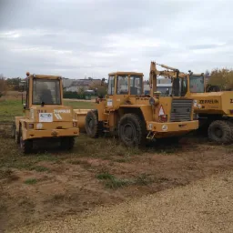 Trzy żółte maszyny budowlane marki Zeppelin stoją na placu budowy z błotnistą ziemią i żwirem, w tle widoczne budynki i drzewa pod pochmurnym niebem.