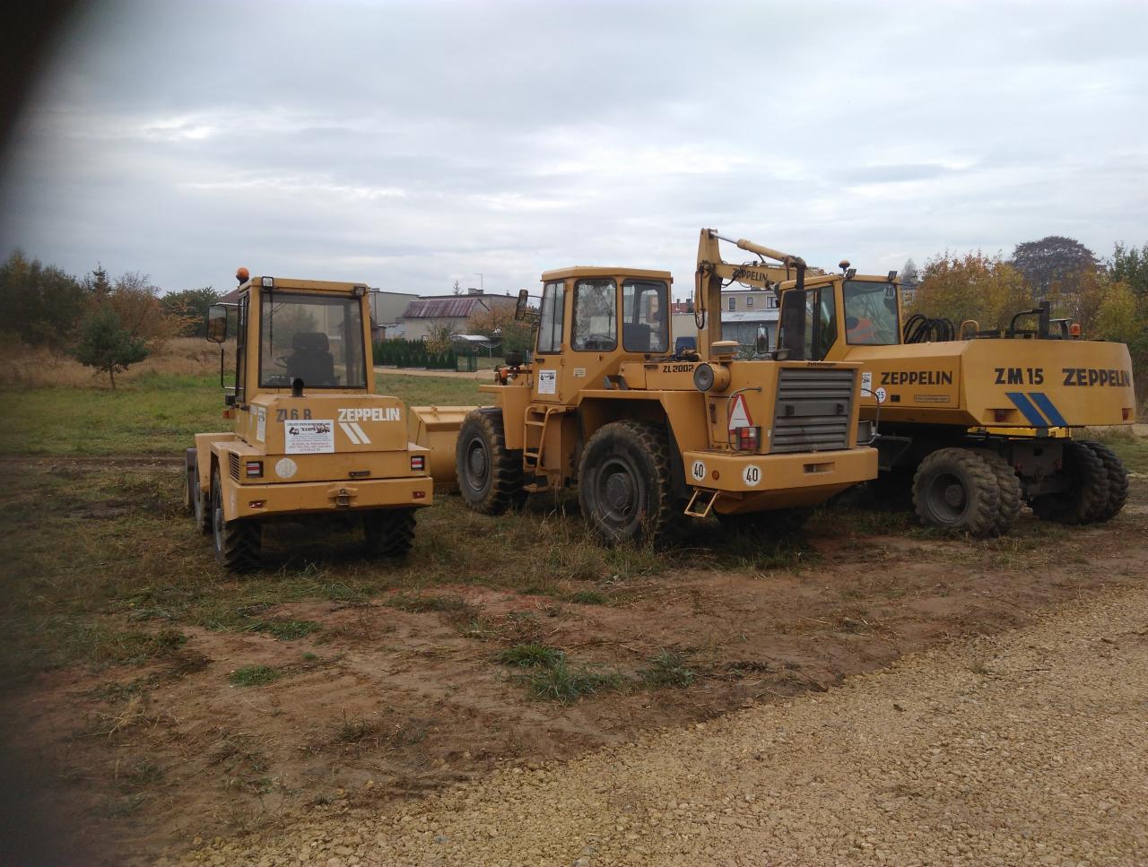 Trzy żółte maszyny budowlane marki Zeppelin stoją na placu budowy z błotnistą ziemią i żwirem, w tle widoczne budynki i drzewa pod pochmurnym niebem.
