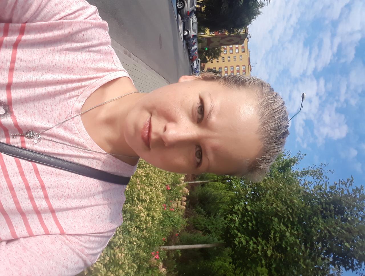 Portret kobiety w różowej koszulce w paski, z naszyjnikiem w kształcie klucza, na tle miejskiego krajobrazu z budynkiem i drzewami.