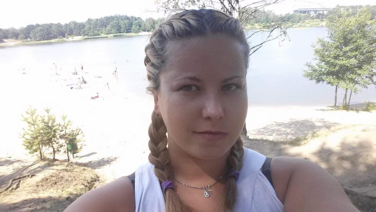 Portret kobiety z warkoczami na tle jeziora i piaszczystej plaży. Uśmiechnięta, z delikatnym makijażem i srebrnym naszyjnikiem. Jasne tło, letni dzień.