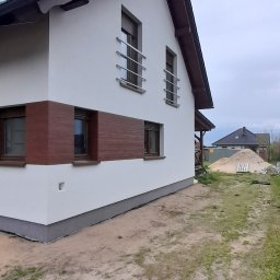 Mar-Bud - Nowoczesny dom jednorodzinny z białymi ścianami, brązowymi akcentami i ciemną dachówką, widoczny z boku na tle pochmurnego nieba i placu budowy.