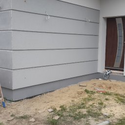 Mar-Bud - Szara elewacja budynku z poziomymi panelami, widoczne zaczepy na ścianie, fragment dachu z szarą dachówką i rynną, teren wokół budynku w trakcie prac wykończeniowych z piaskiem i cegłami.