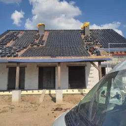 Dom w trakcie krycia dachu czarną dachówką, widoczne kominy zabezpieczone żółtą folią, na pierwszym planie fragment samochodu dostawczego.