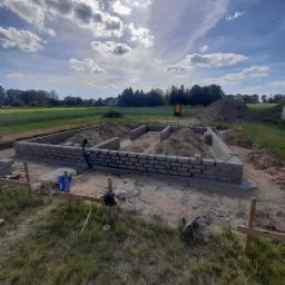 Fundamenty domu w trakcie murowania z bloczków betonowych, widoczne przygotowane wykopy, teren budowy z trawą i narzędziami, słoneczny dzień z chmurami w tle.