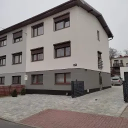 Dwukondygnacyjny dom z elewacją w dwóch odcieniach szarości i brązowymi roletami, numer domu 27, widok z poziomu ulicy w pochmurny dzień.