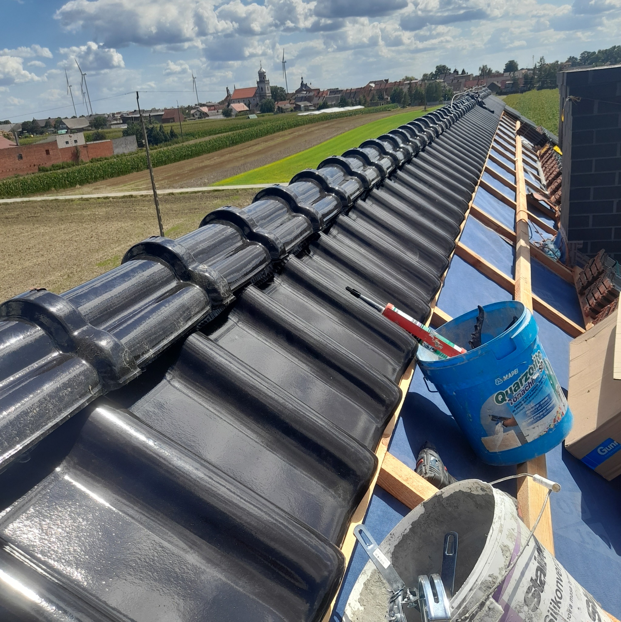 Częściowo pokryty dach z błyszczącej, czarnej dachówki ceramicznej, widoczne narzędzia i wiaderka z materiałami budowlanymi, w tle wieś z wiatrakami.